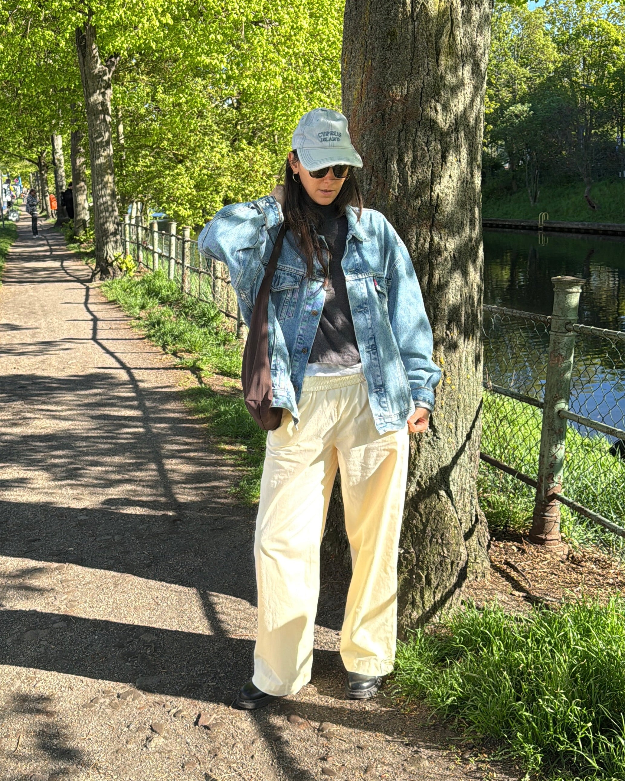 A person standing outdoors on a tree-lined path next to a body of water, wearing a light blue denim jacket, cream wide-leg pants, a light blue cap, and dark sunglasses.