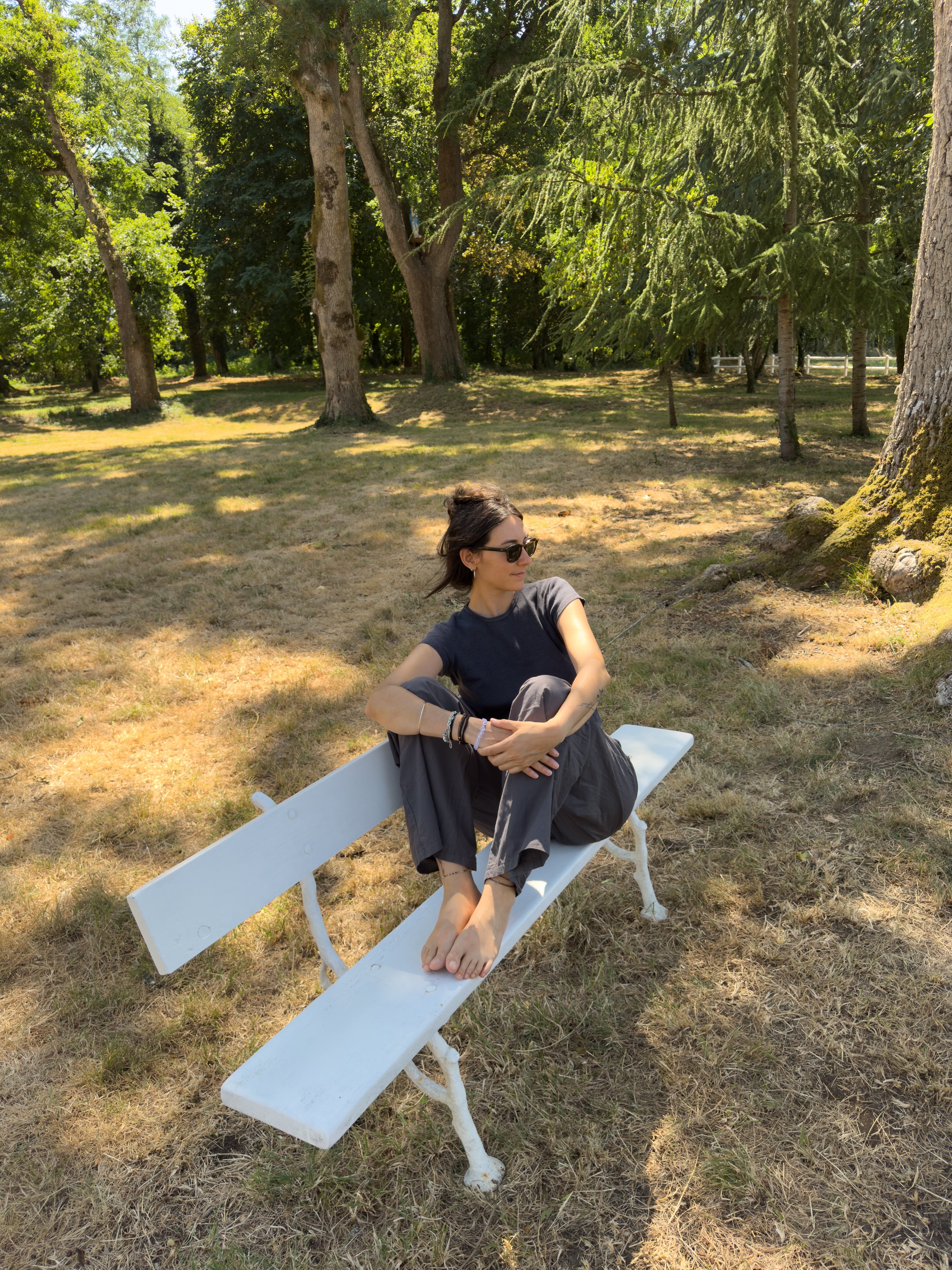 A woman sits barefoot on a white bench in a grassy park area with trees in the background.